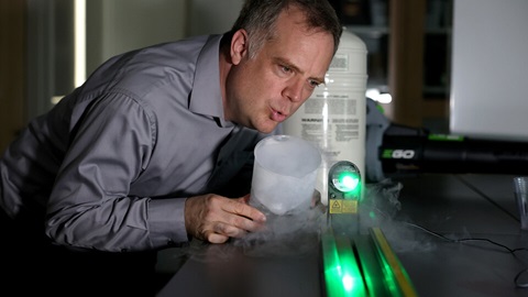 In a lab setting, University of Nebraska at Kearney Physics Professor Jeremy Armstrong leans into an experiment showing gas and a bright green light.
