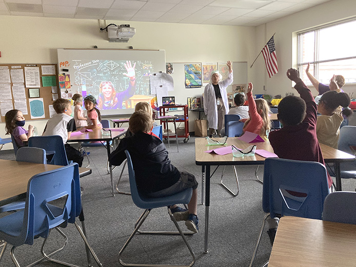 In an elementary school classroom, a Zoom session is projected on a big screen with Professor Sko (a fun science educator) waving to the 12 students and 2 teachers who are waving back.