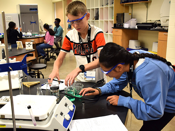 a boy and a girl work with liquids in science flasks while other students gather at a table in a lab