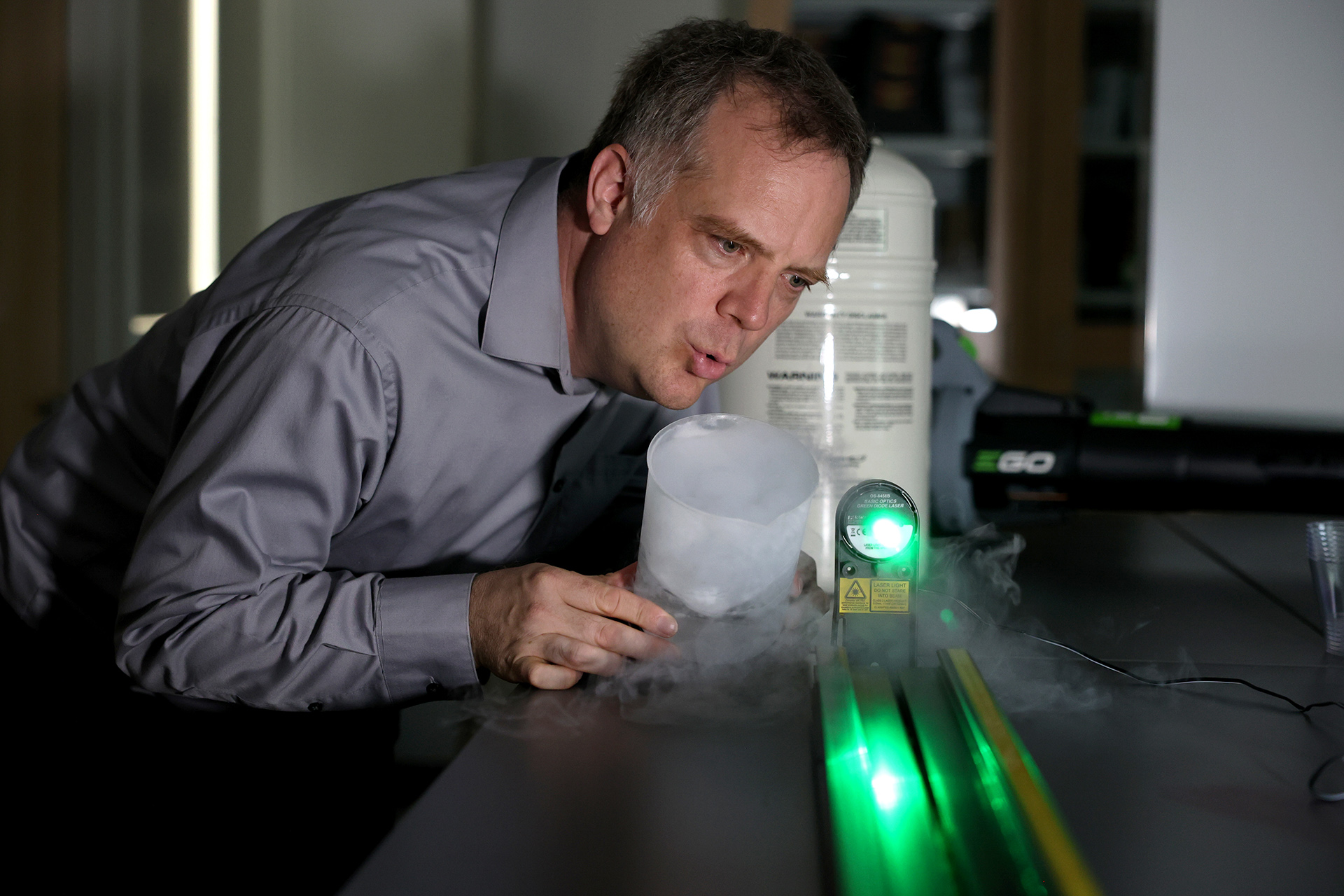 In a lab setting, University of Nebraska at Kearney Physics Professor Jeremy Armstrong leans into an experiment showing gas fumes and a bright green light.